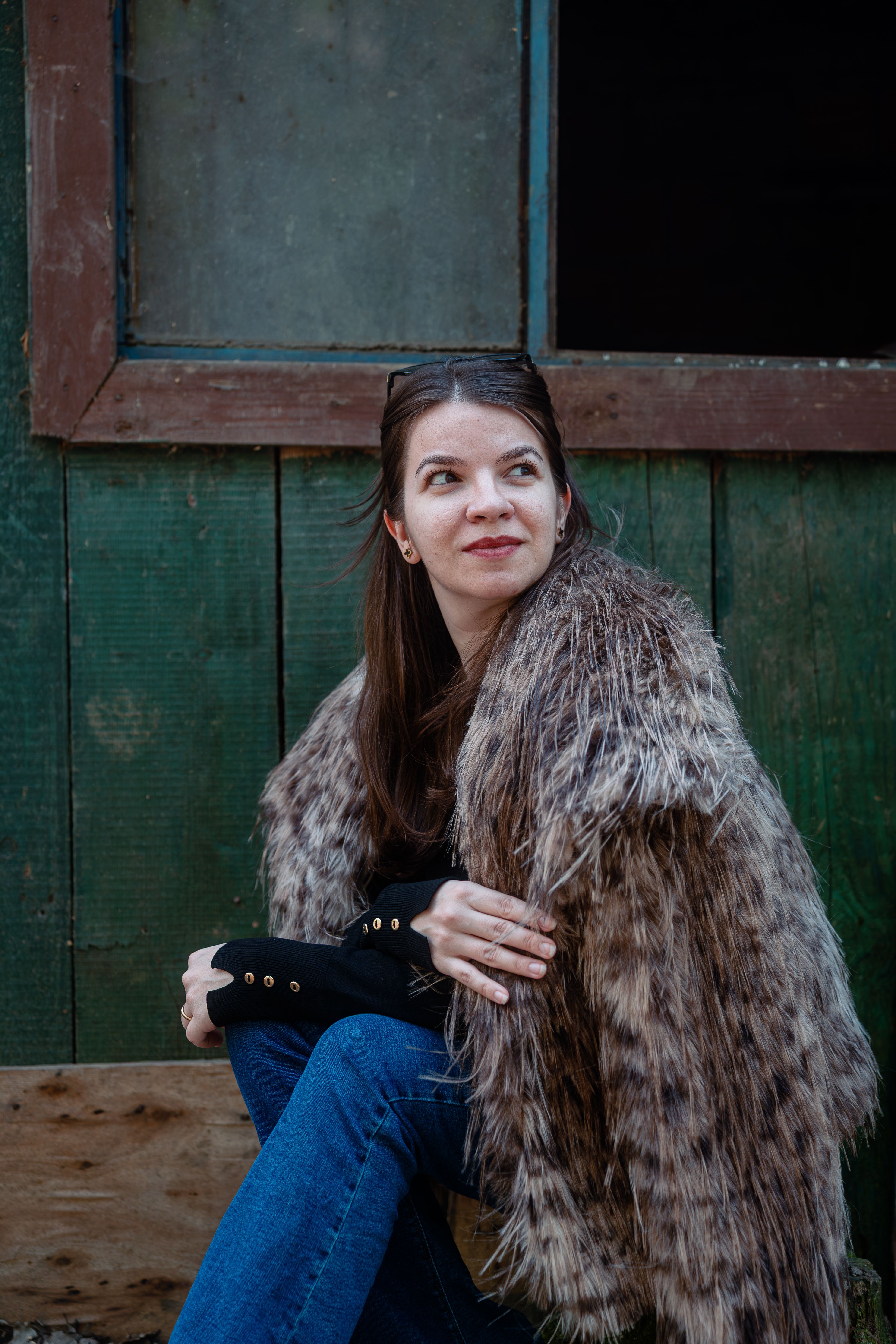 Portrait of a woman in a faux fur coat sitting against a rustic green wall.