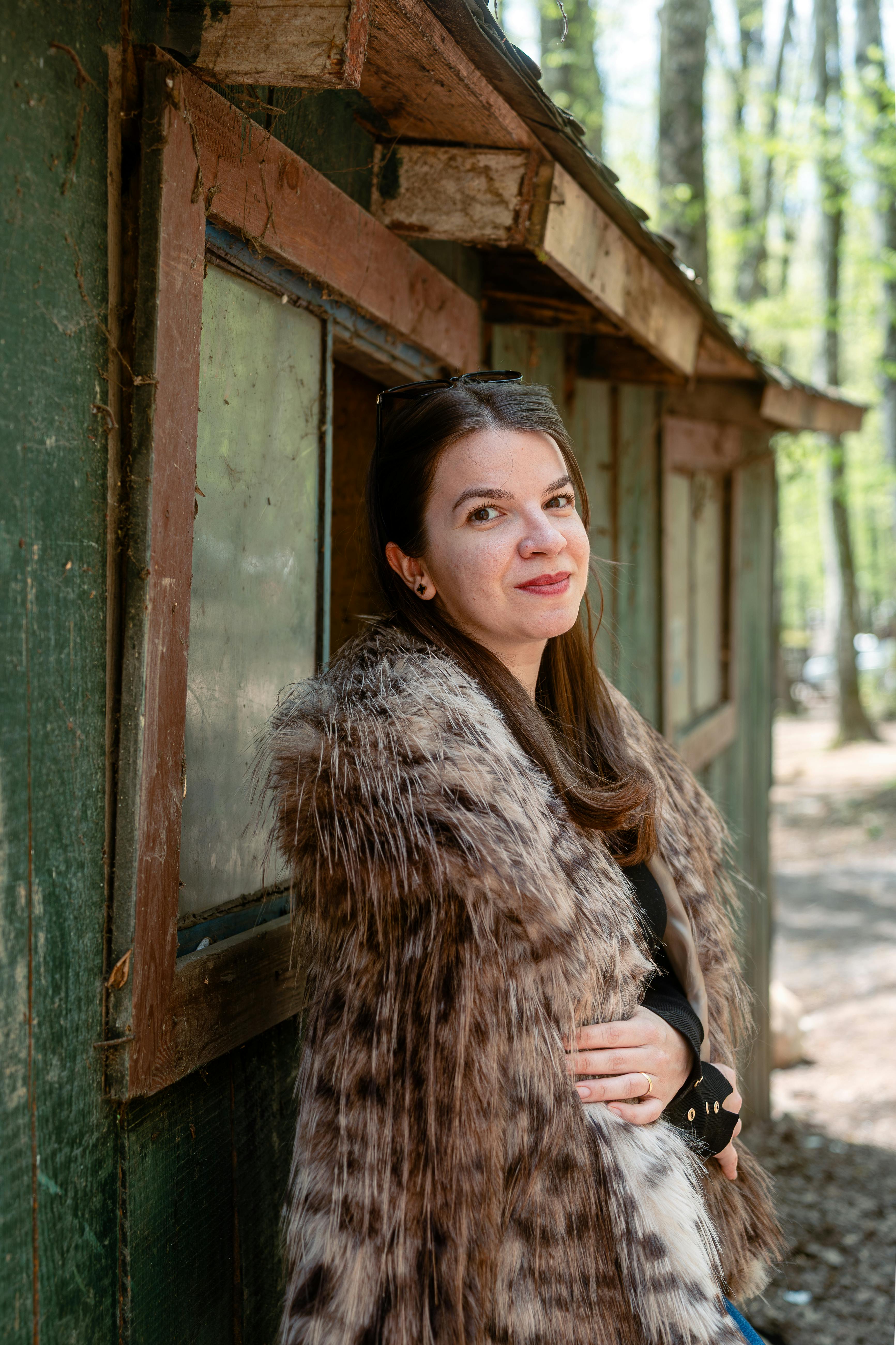 Stylish woman in fur coat smiling by a rustic cabin in a forest setting.