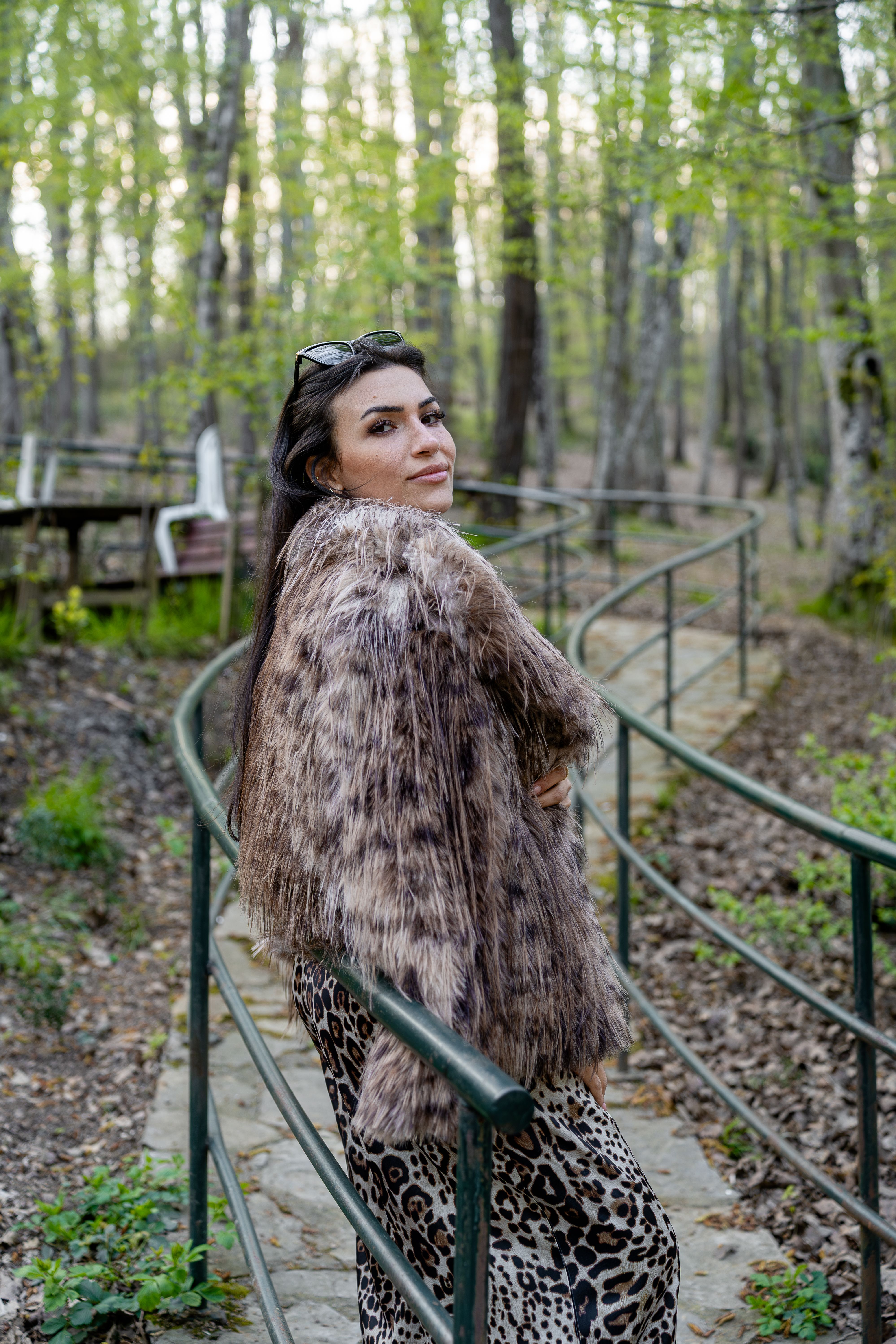 Stylish woman in faux fur coat walks through a serene woodland path, embracing nature.
