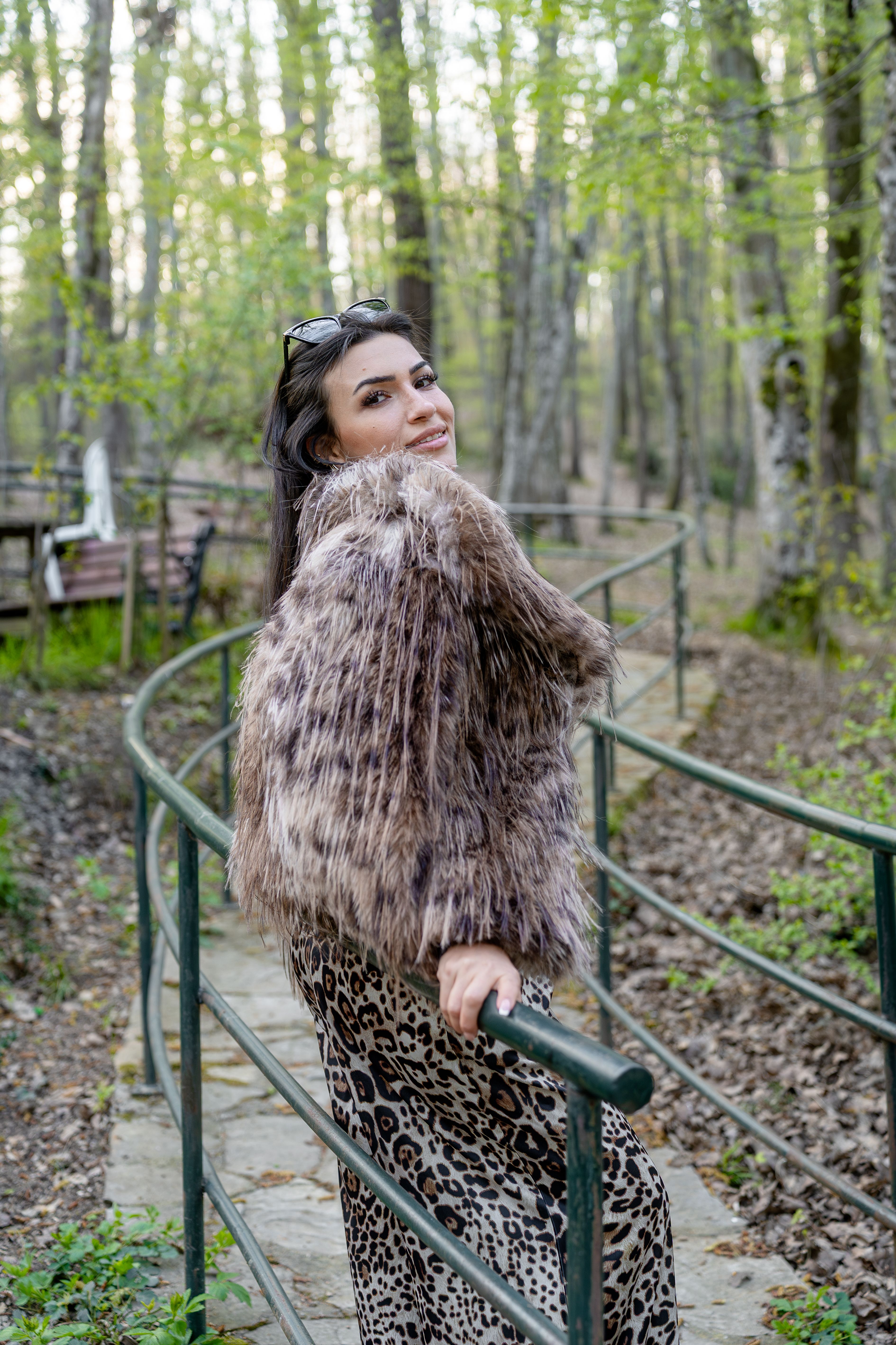 Fashionably dressed woman in fur coat smiles while strolling through a lush spring forest.