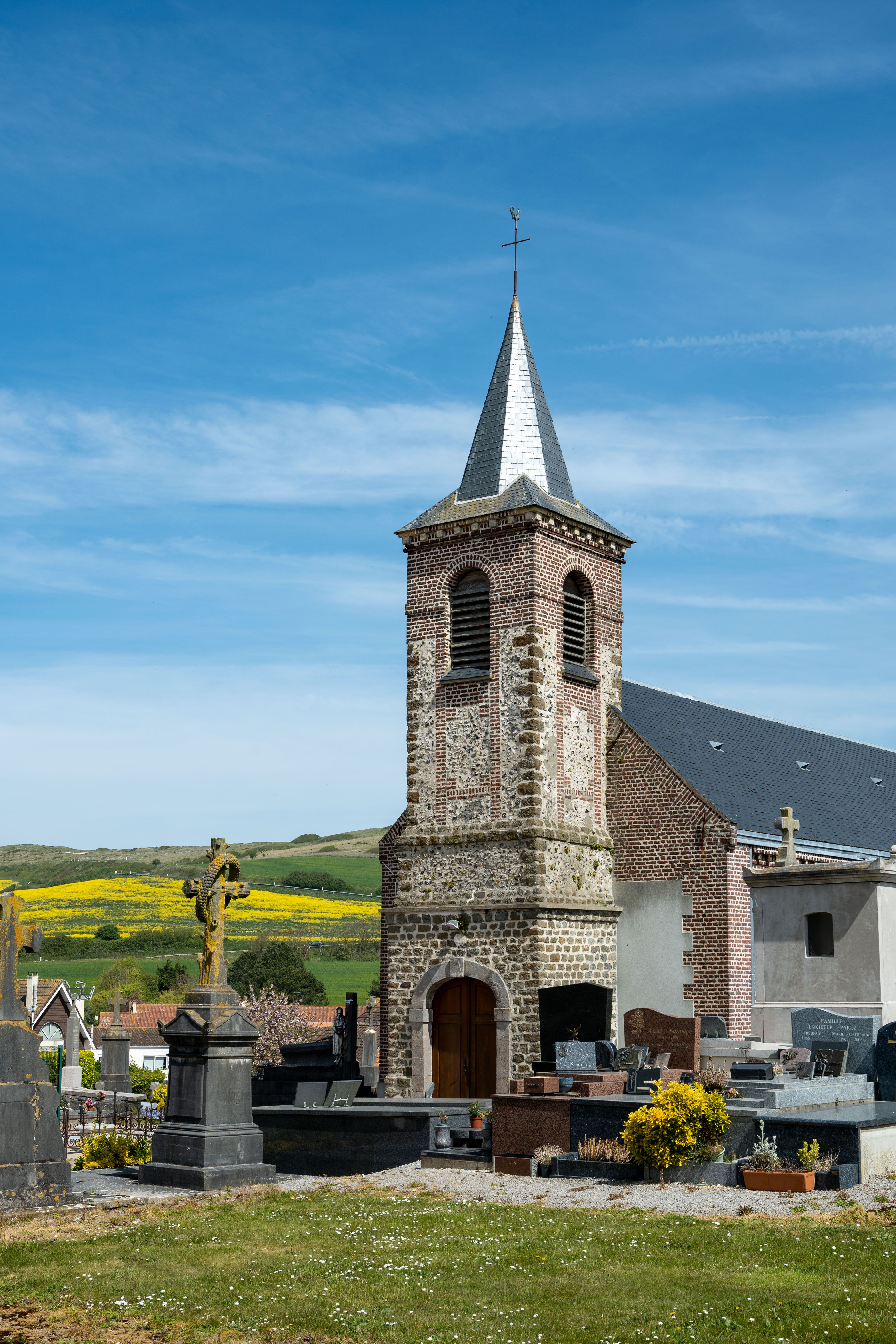 A captivating image of a historic church tower in Escalles, France, surrounded by lush greenery and a graveyard.
