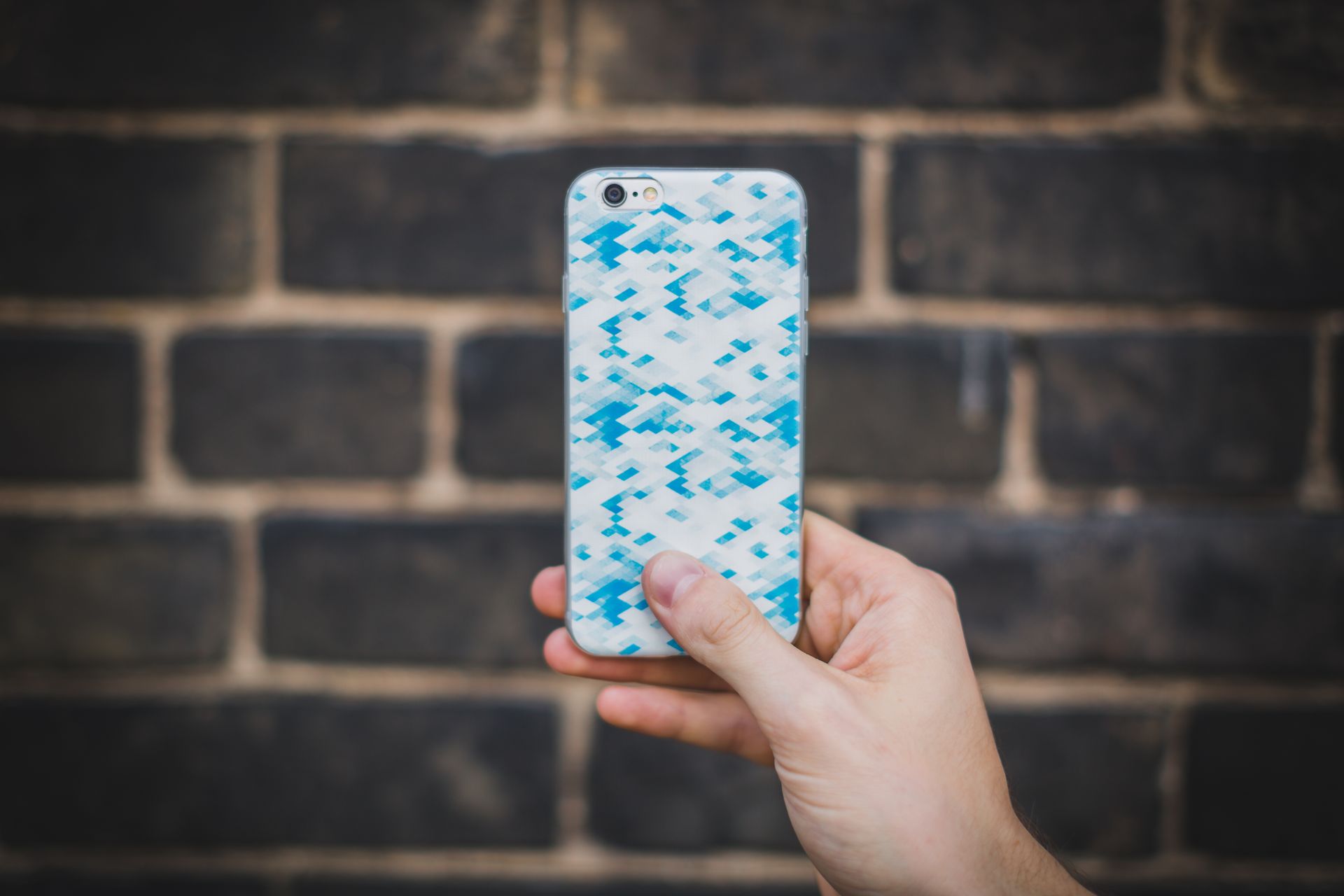 A hand holds a smartphone with a blue patterned case in front of a textured brick wall.