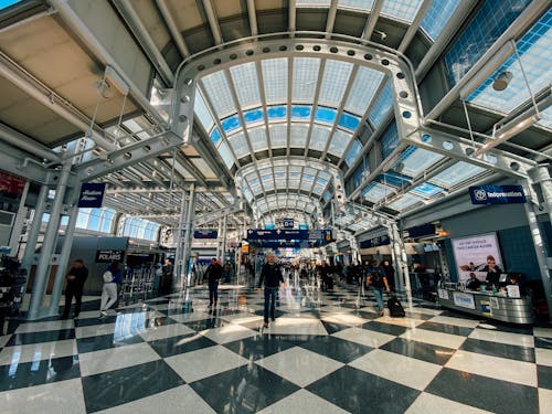 Contemporary style building interior with many symmetric lamps on ceiling and metal beams above tile floor with people strolling in spacious hall