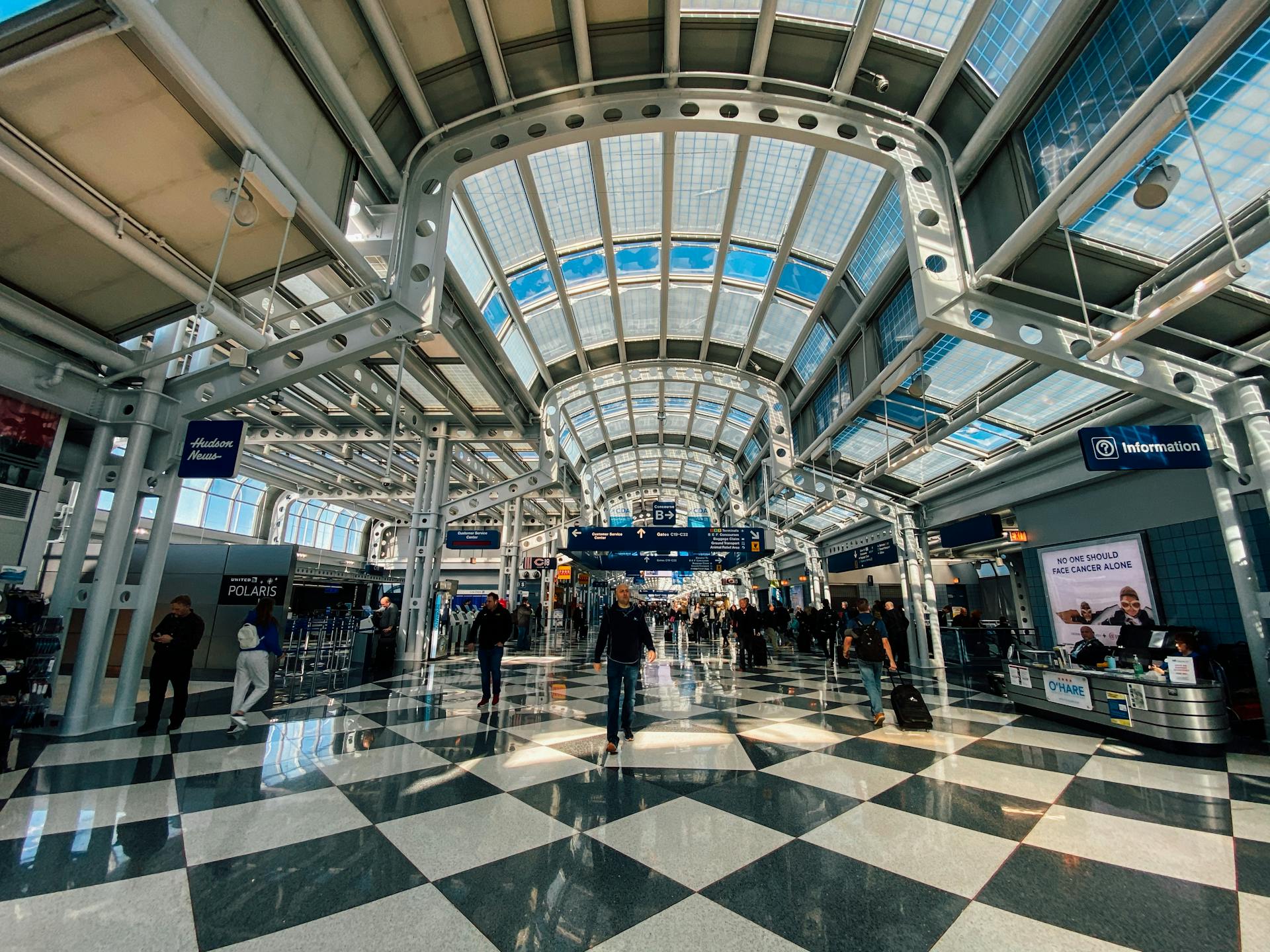 Contemporary style building interior with many symmetric lamps on ceiling and metal beams above tile floor with people strolling in spacious hall