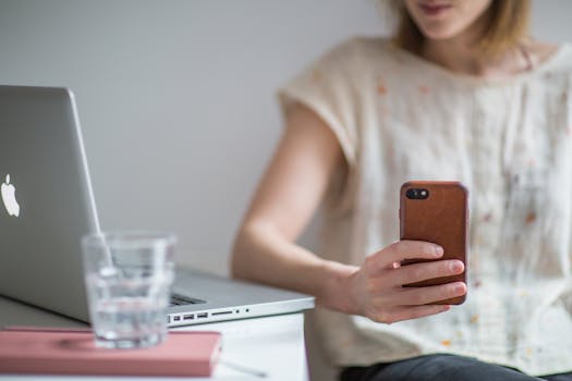 Young woman using smartphone next to a laptop on a desk indoors.