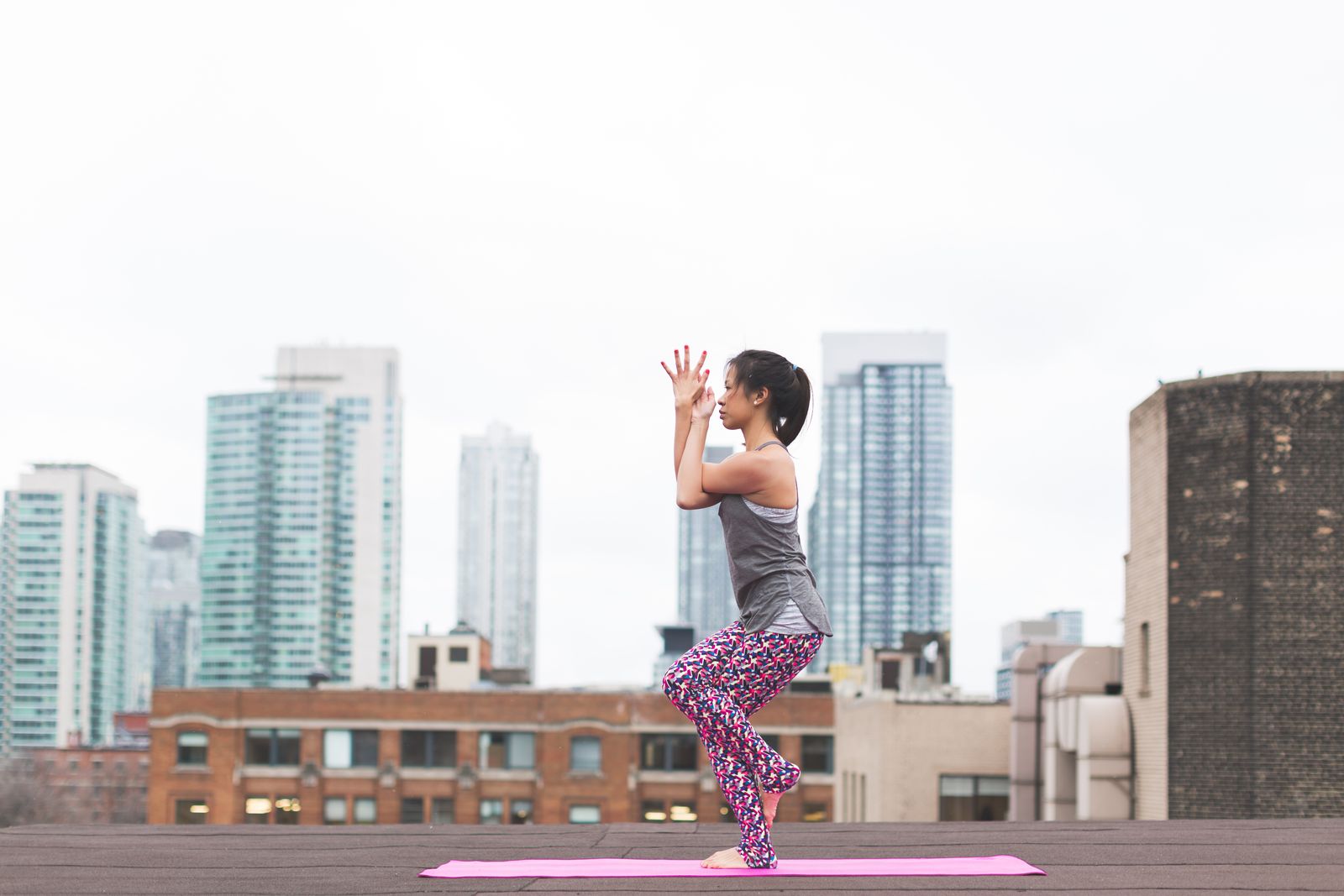 Woman practicing eagle arms yoga pose on a rooftop with a city skyline behind her