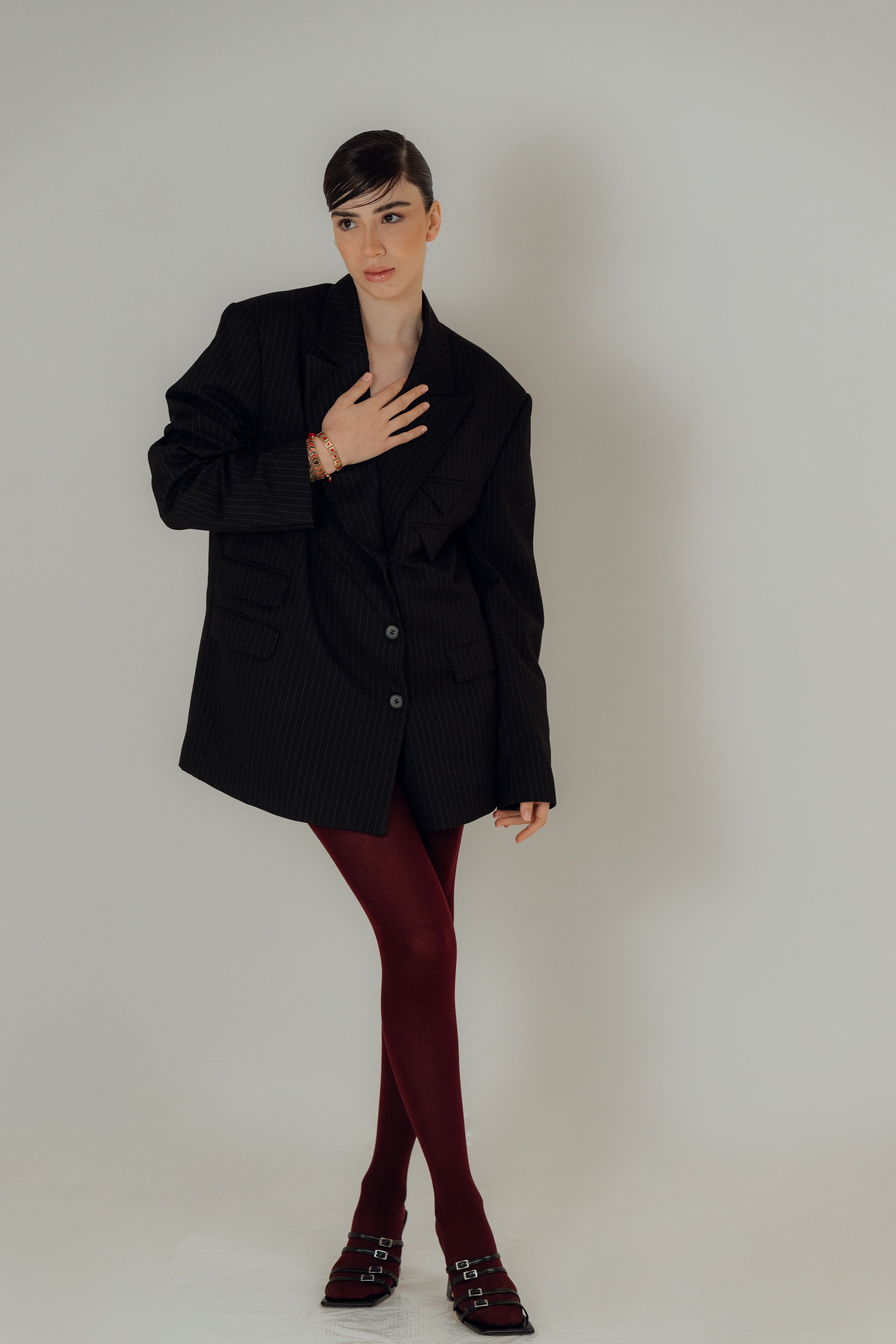 Mulher estilosa vestindo um blazer preto oversized e leggings bordô, posando de forma confiante contra um fundo neutro.