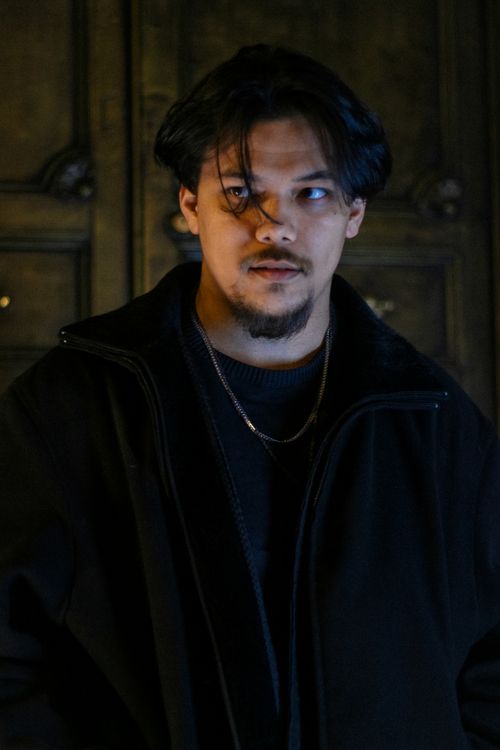 Free Portrait of a young man in a black coat standing indoors against an ornate background. Stock Photo