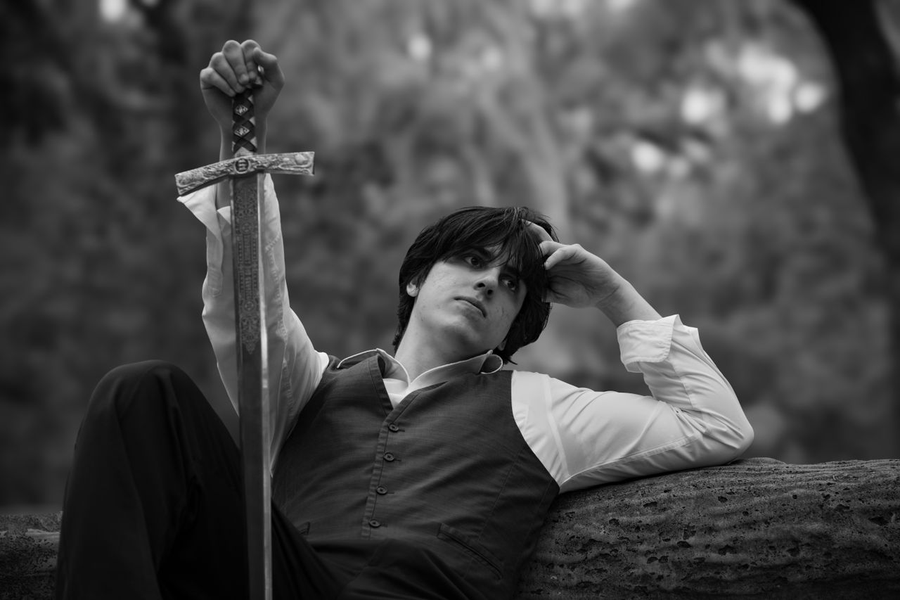 a pensive young man in medieval attire holding a sword set in nature