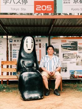 A man seated with No-Face statue at a themed bus stop in Japan.