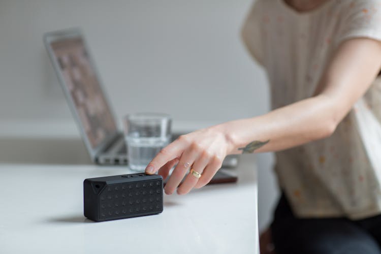 Woman Reaching For Black Bluetooth Speaker On Top Of White Table