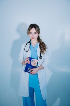 A young female doctor in scrubs and lab coat holding a medical book, standing indoors.