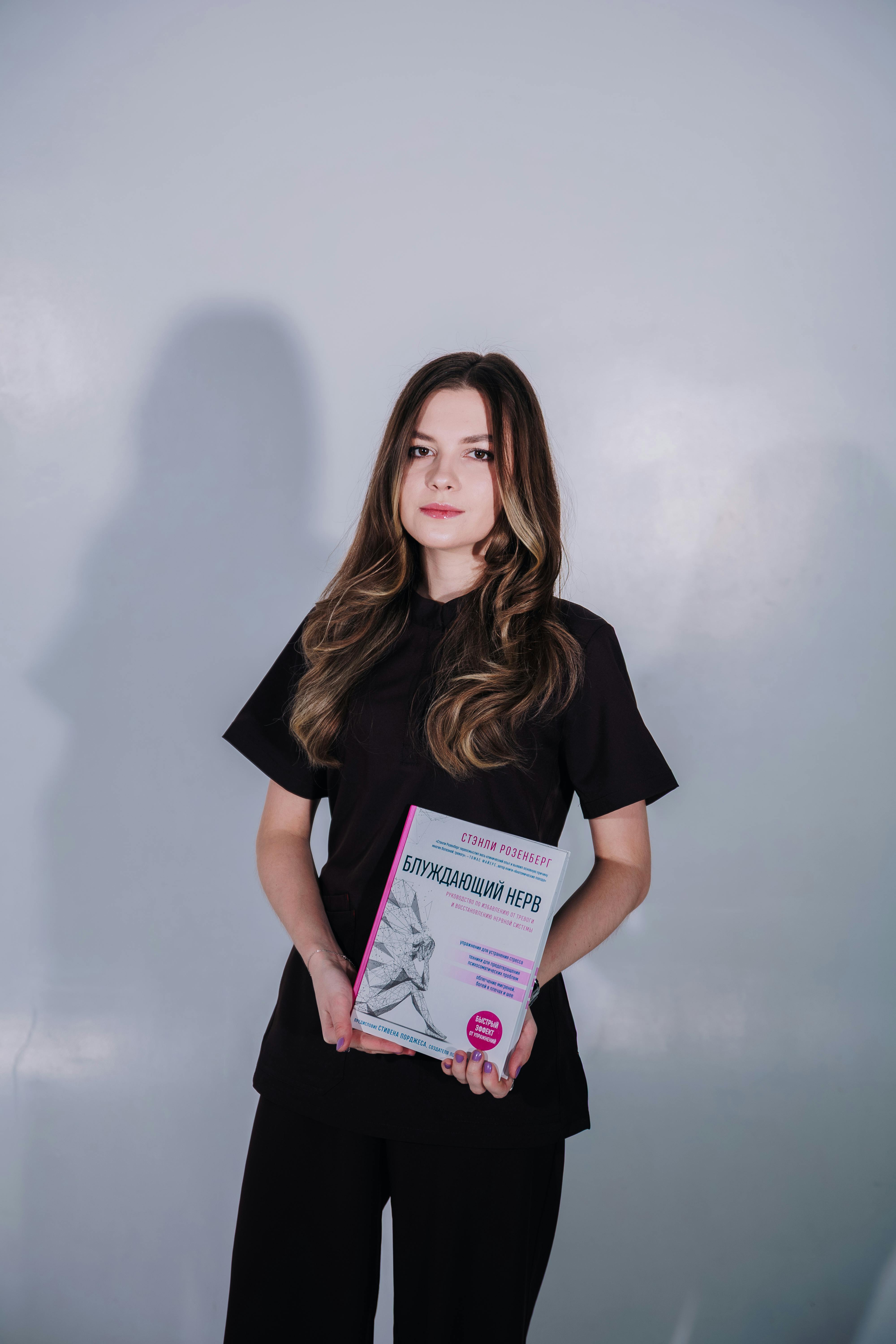 A young woman standing indoors holding a book with a light gray background.