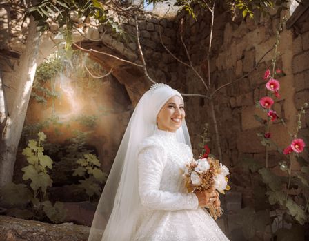 A beautiful bride in a white dress and veil holds a bouquet, smiling in a rustic outdoor setting.