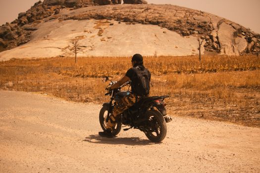 A lone motorcyclist rides through a barren, rocky desert landscape under the sun.