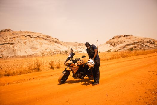 A man inspects his motorcycle on a sandy road amid rocky desert scenery under the bright sun.