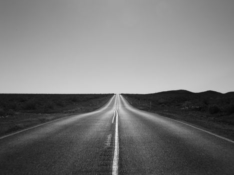A black and white photograph of a straight desert road leading into the horizon.
