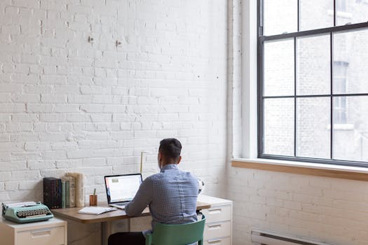 Adult male working remotely on a laptop in a well-lit home office with a large window.