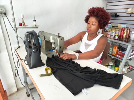 An attentive seamstress working on a black fabric using a mechanical sewing machine in a small workshop.