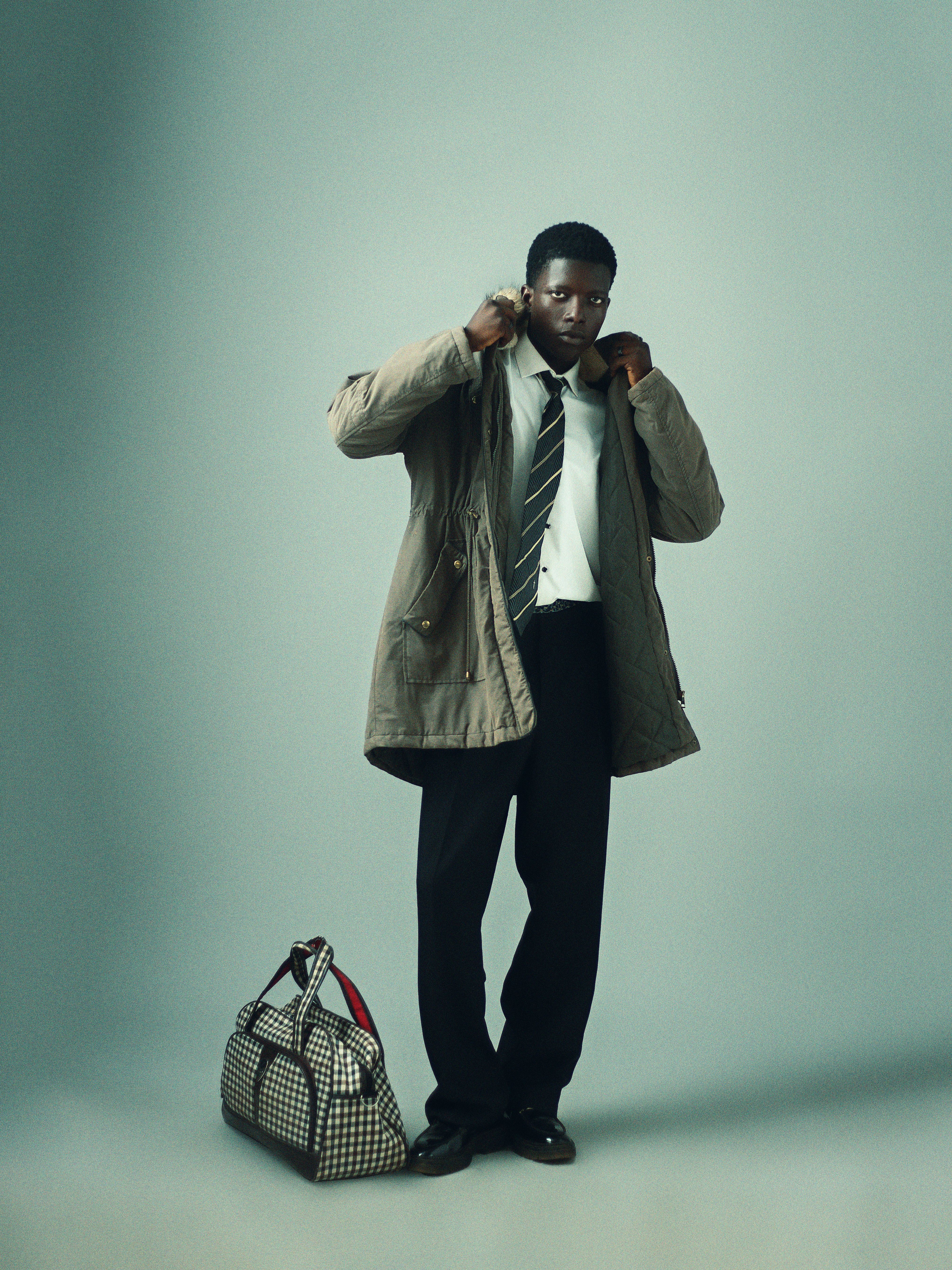 Stylish young adult in formal attire adjusting coat, standing with travel bag.