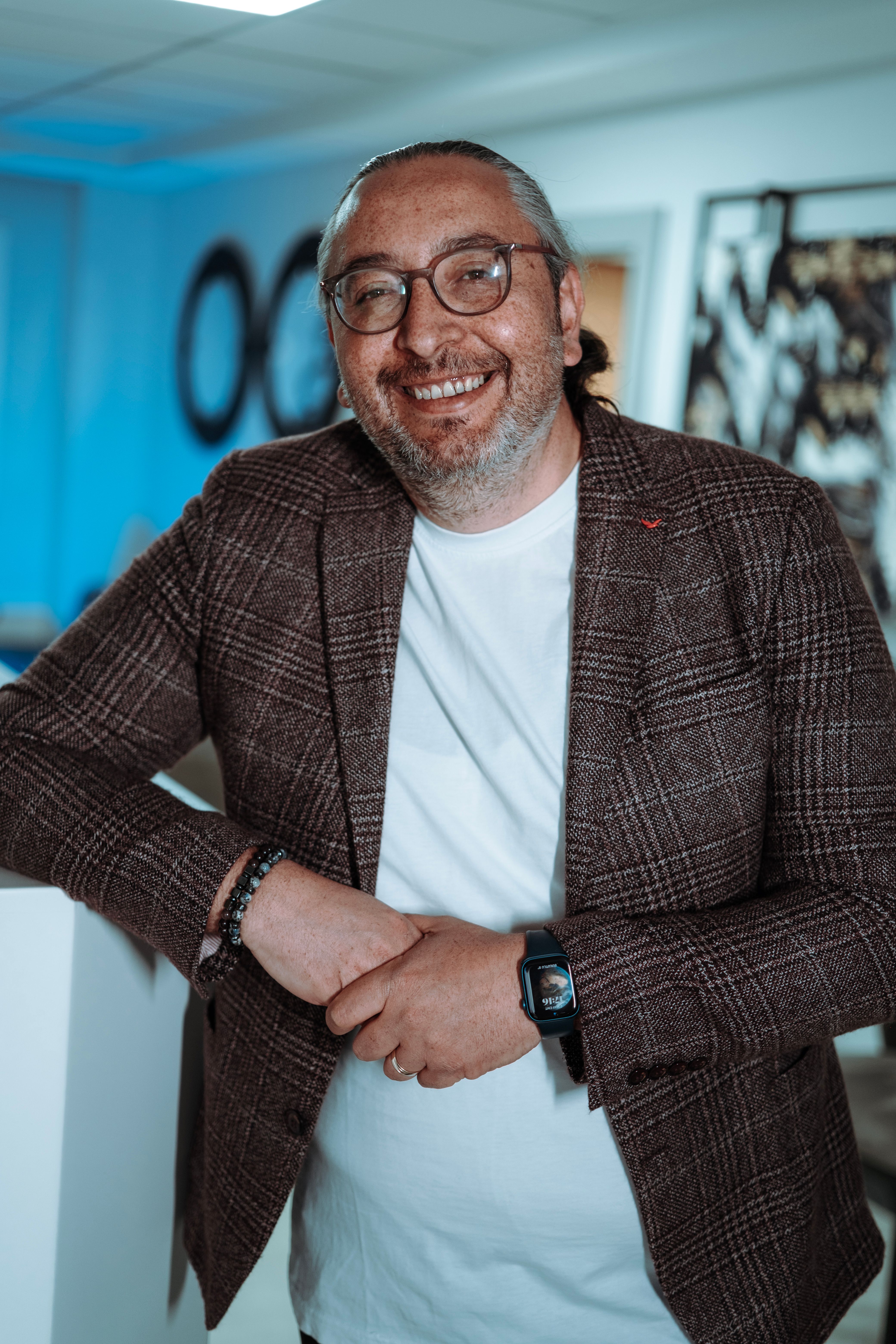 Happy adult man with glasses in a modern office setting, leaning on a desk and smiling.