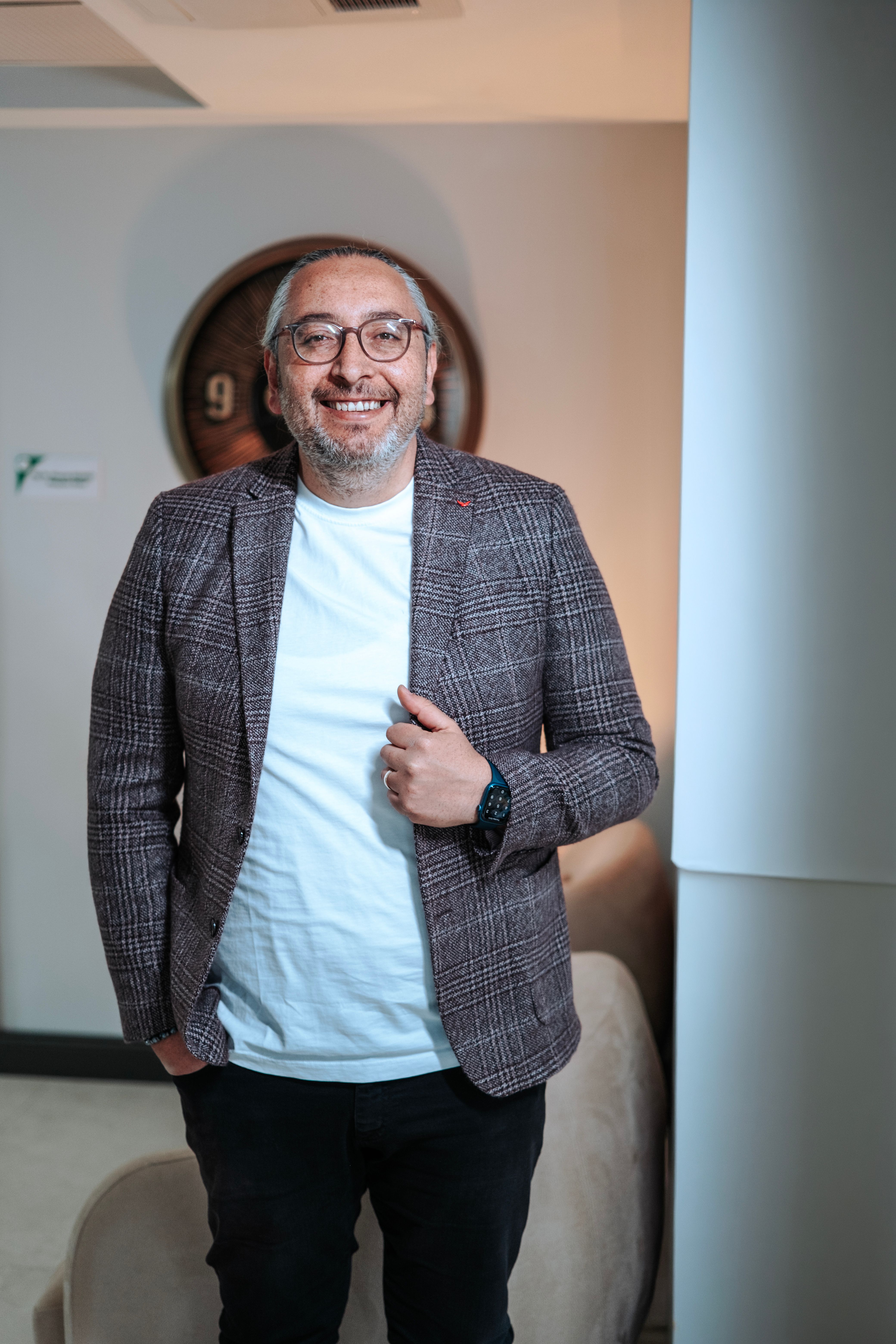 Portrait of a smiling adult male wearing glasses and a stylish plaid jacket in an indoor setting.