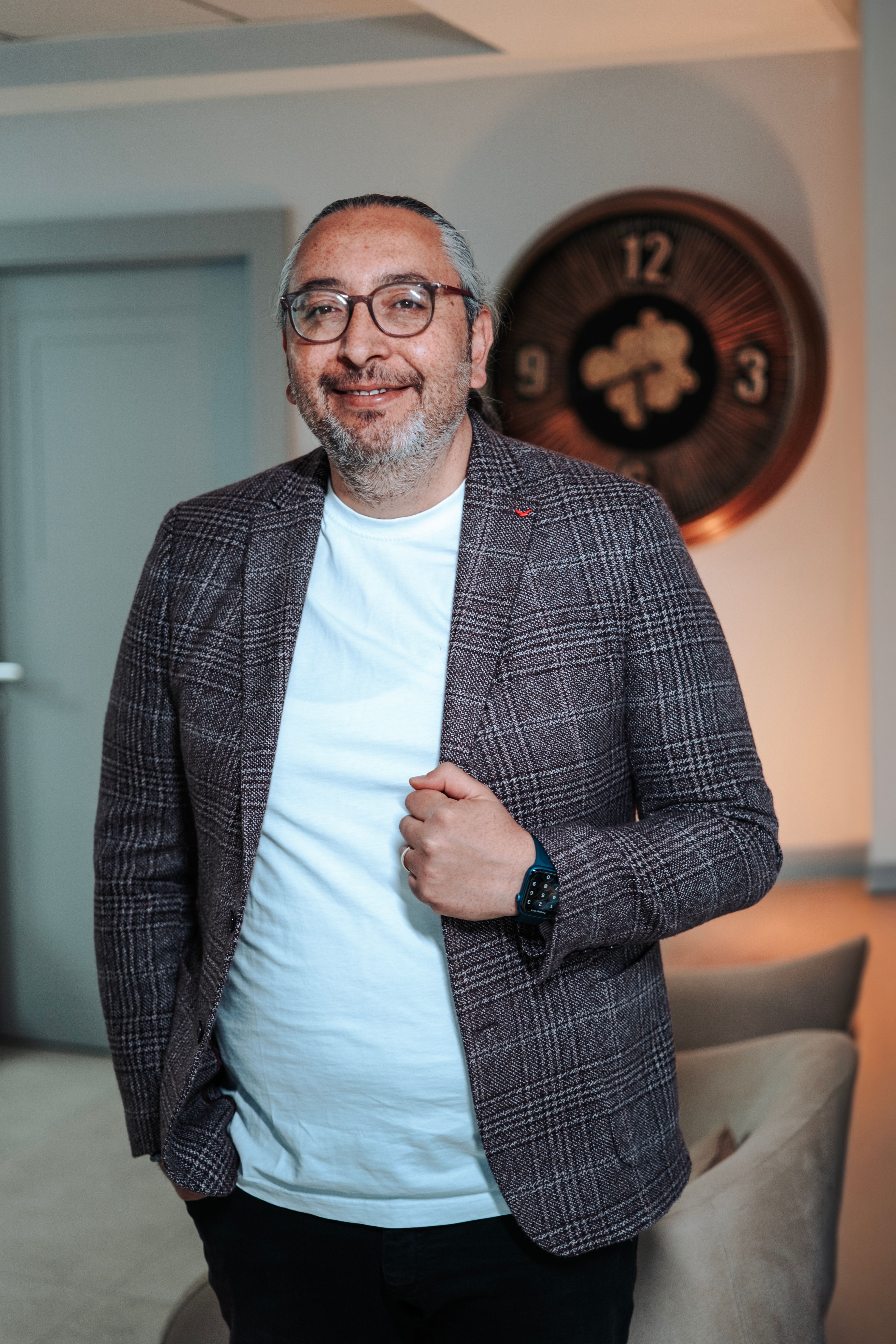 Smiling businessman with glasses and blazer standing confidently in office setting with stylish decor.