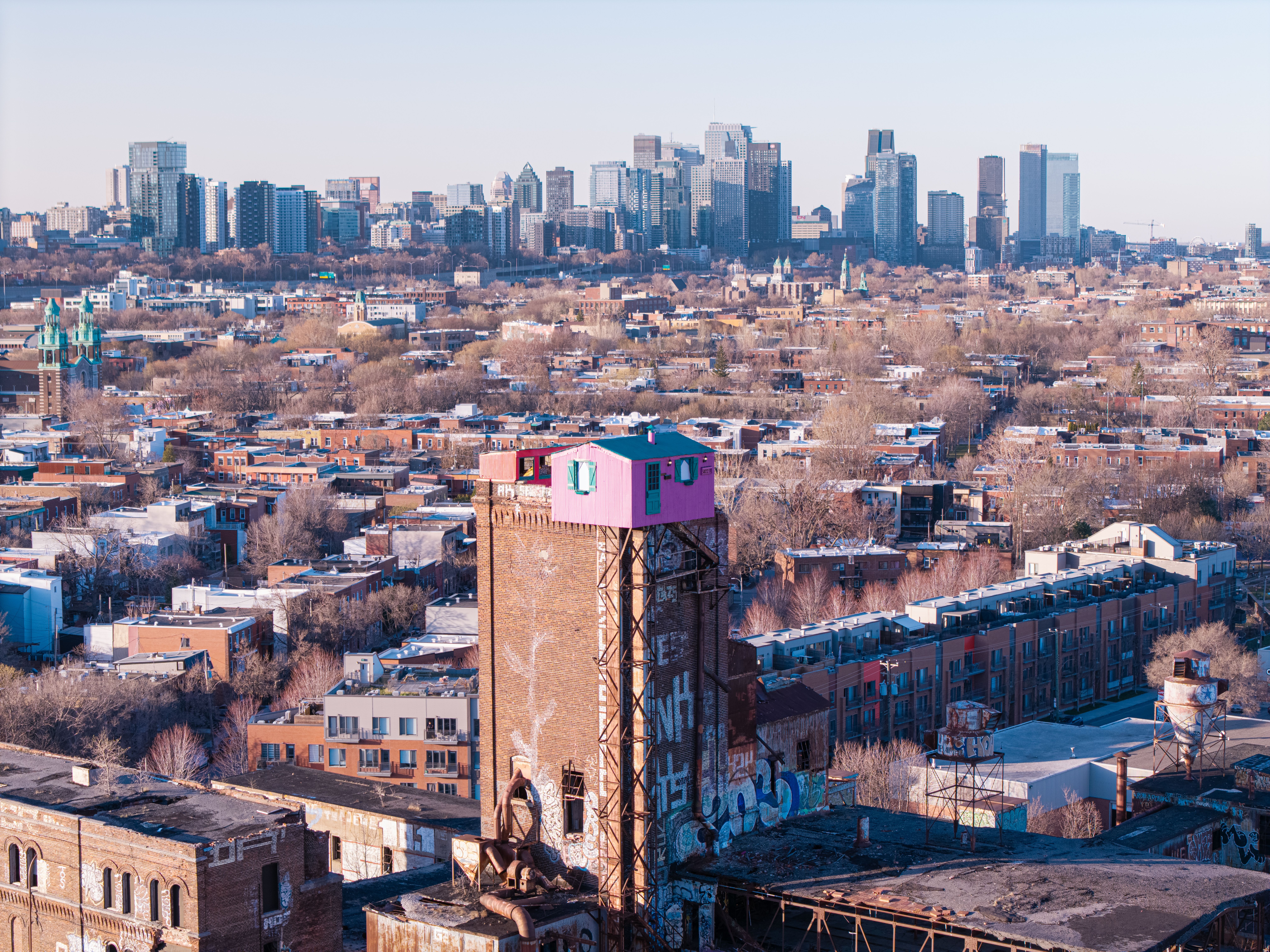 Aerial view capturing Saint-Henri's industrial landscape with Montreal's skyline, highlighting a vibrant pink rooftop.