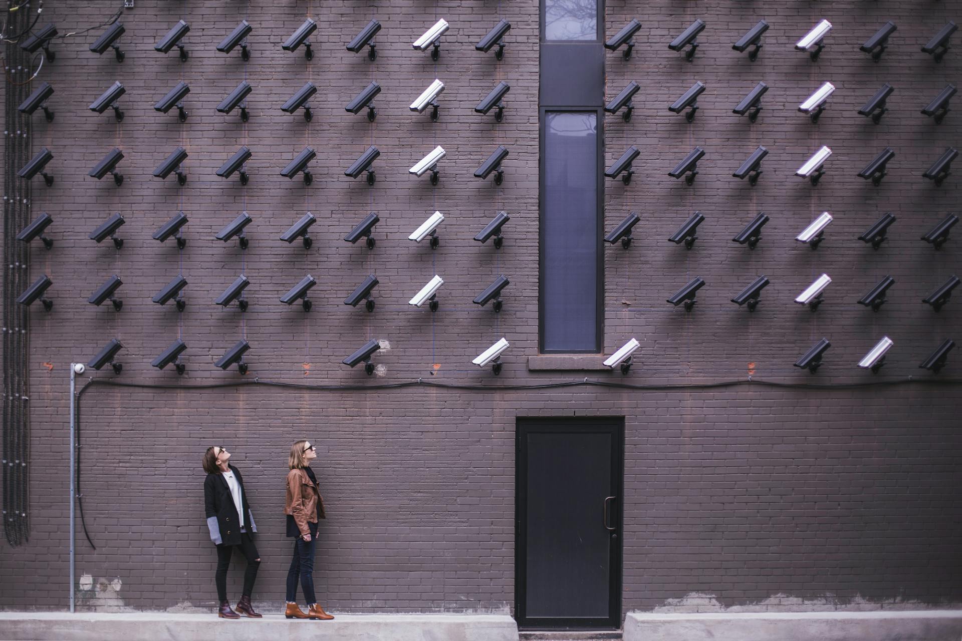 Women observing a wall covered with numerous security cameras in an urban setting, showcasing modern surveillance.