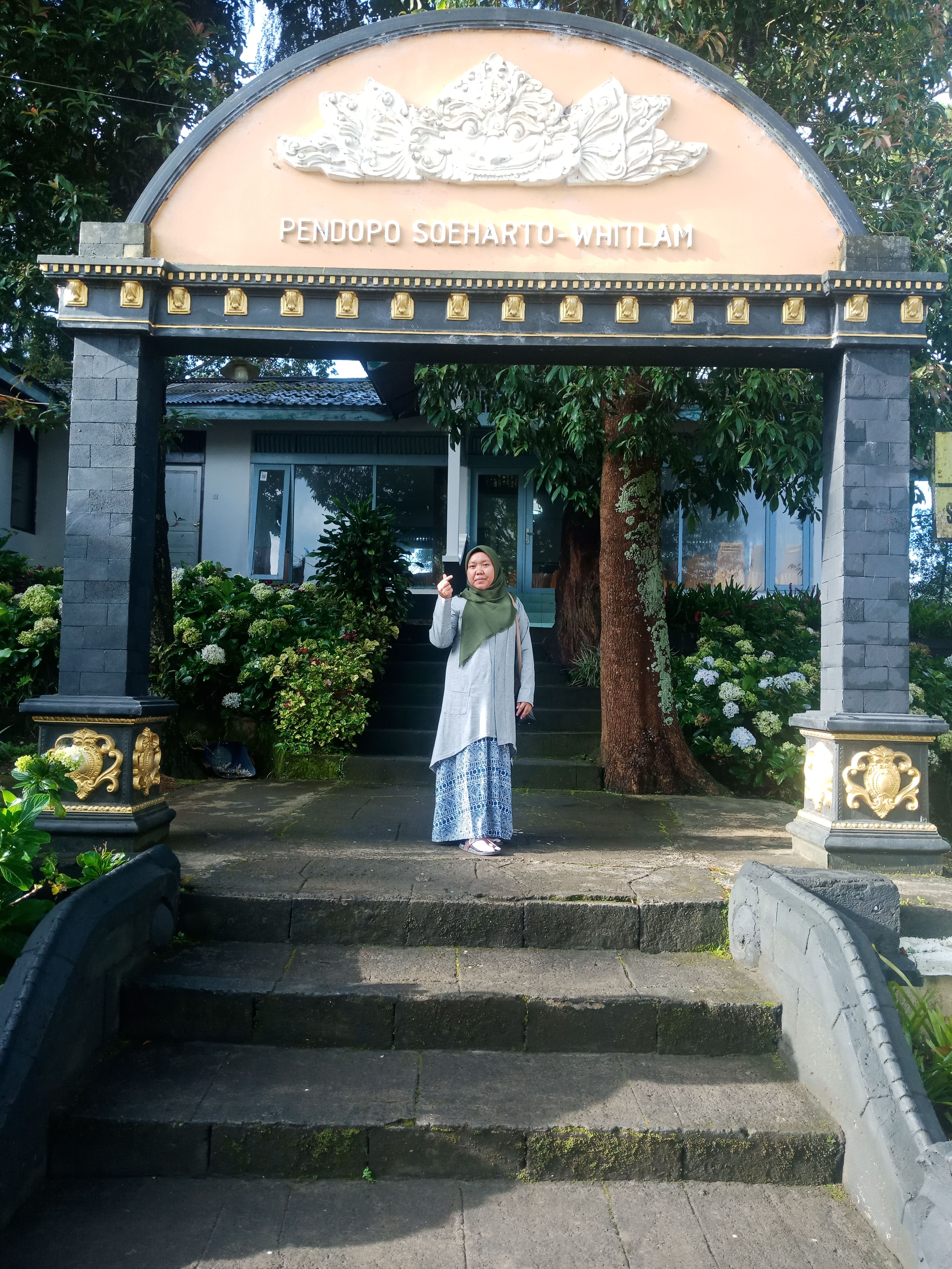 A woman poses beneath the ornate Pendopo Soeharto Whitlam gate amidst lush greenery.