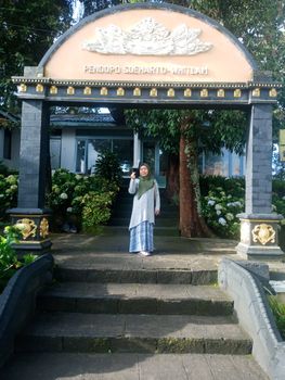 A woman poses beneath the ornate Pendopo Soeharto Whitlam gate amidst lush greenery. - Fotografer Sidoarjo