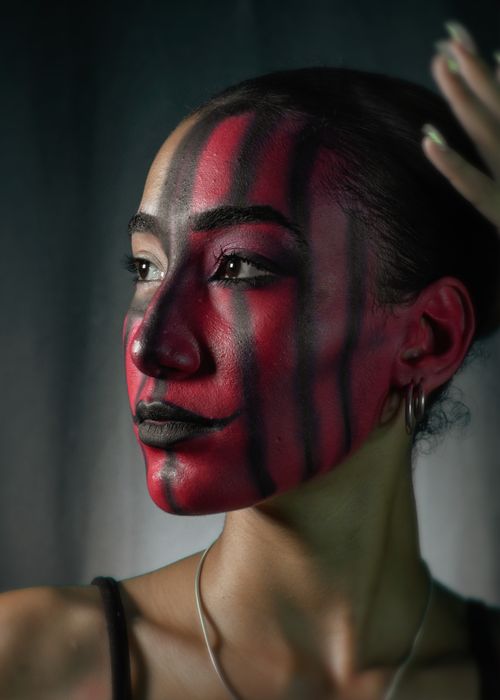 Free A striking artistic portrait featuring bold red and black face paint on a young woman, showcasing creative makeup design. Stock Photo