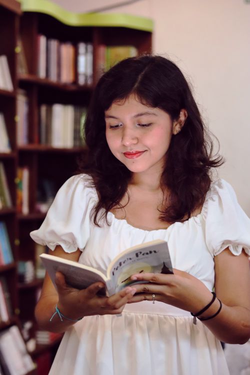 Free A young Hispanic woman in a white dress enjoys reading a book in a cozy bookstore setting. Stock Photo