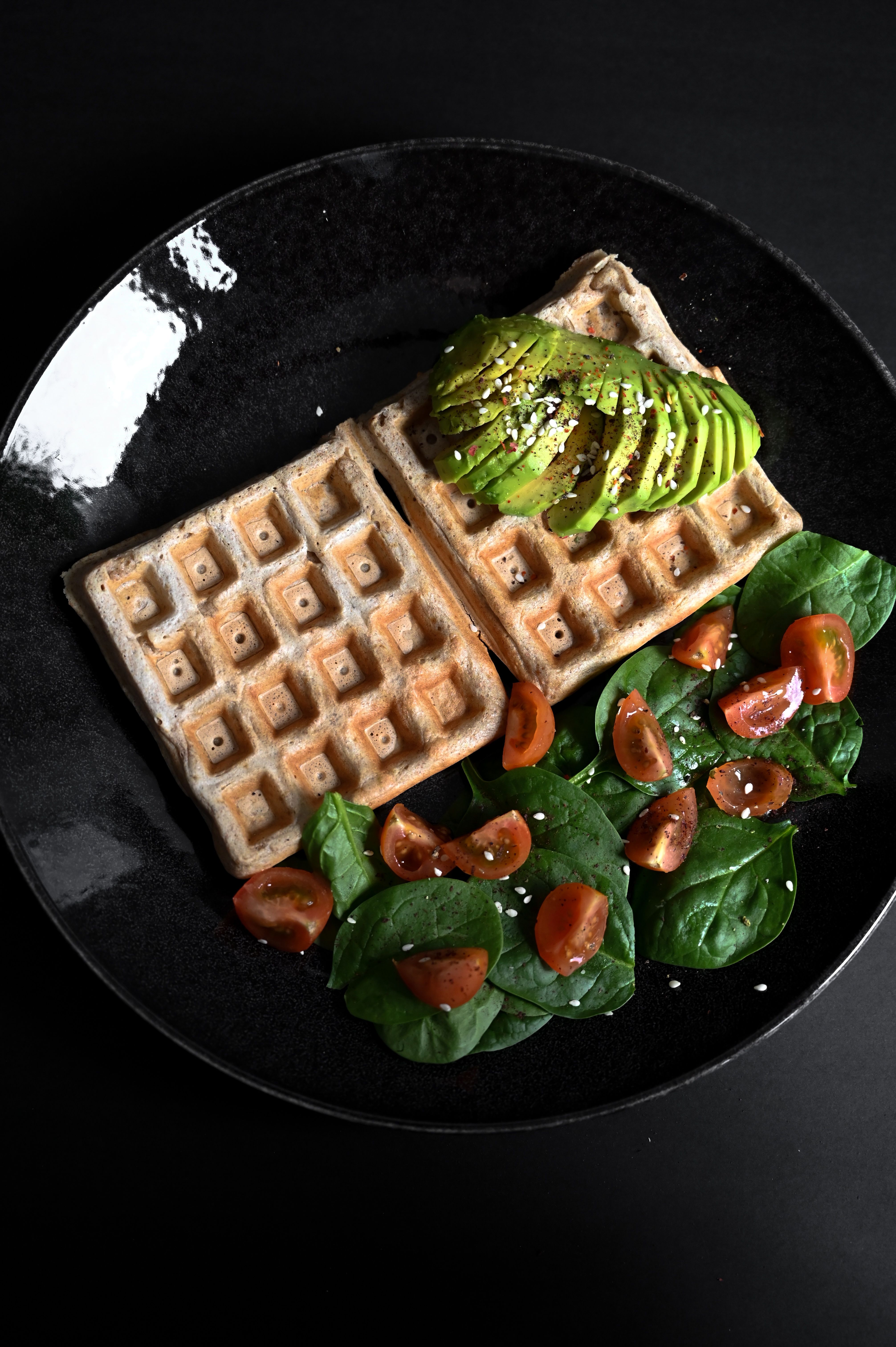Delicious waffle topped with fresh avocado, spinach, and cherry tomatoes on a black plate.