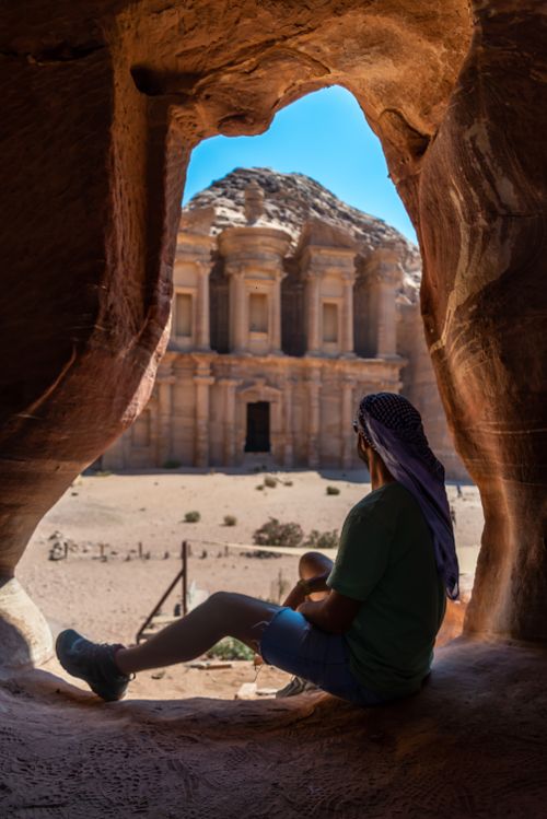 Free Traveler sitting in a cave overlooking Petra's iconic ancient architecture under a bright blue sky. Stock Photo