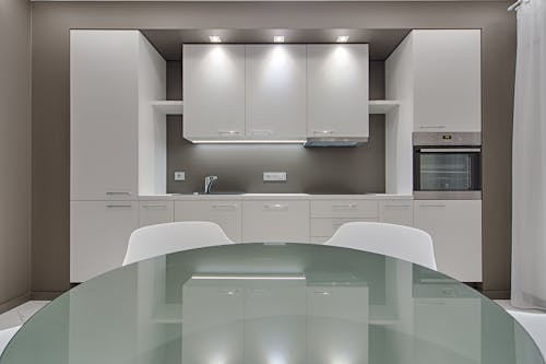 Design of kitchen with white cabinets and contemporary appliances with glass table and white chairs in apartment