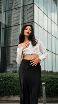 Confident woman in elegant outfit posing against modern skyscraper background, urban fashion vibe.
