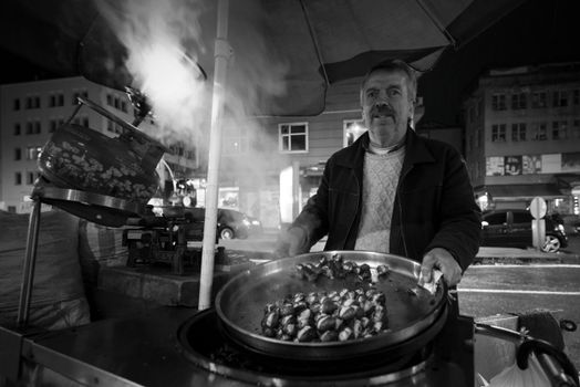 A street vendor grills chestnuts outdoors at night. The scene is atmospheric with steam and city lights.