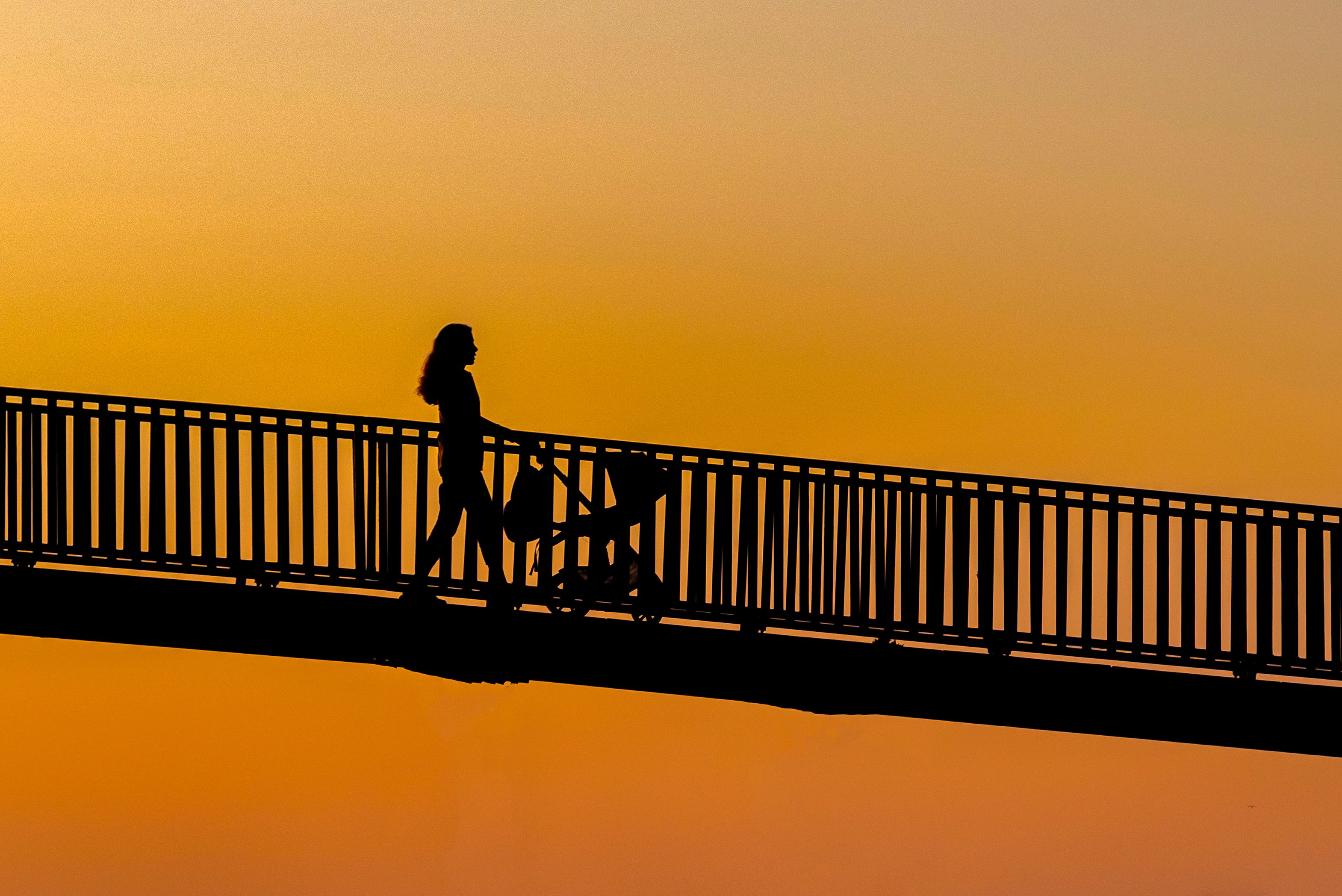 A silhouette of a woman pushing a stroller on a bridge against a vibrant sunset background. Perfect for themes of motherhood or tranquility.