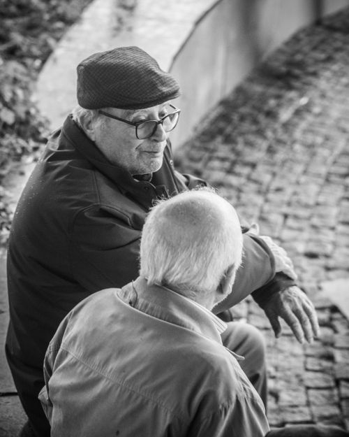 Free Black and white photo of two senior men sitting and talking on a cobblestone street. Stock Photo