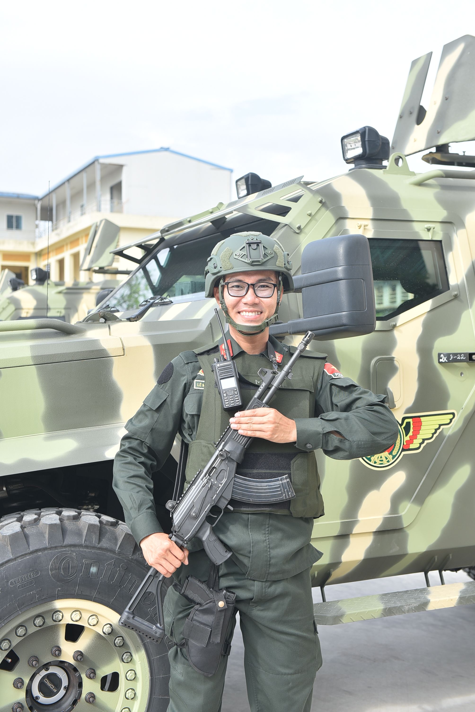 Smiling soldier in uniform posing with camouflaged armored vehicle outdoors.