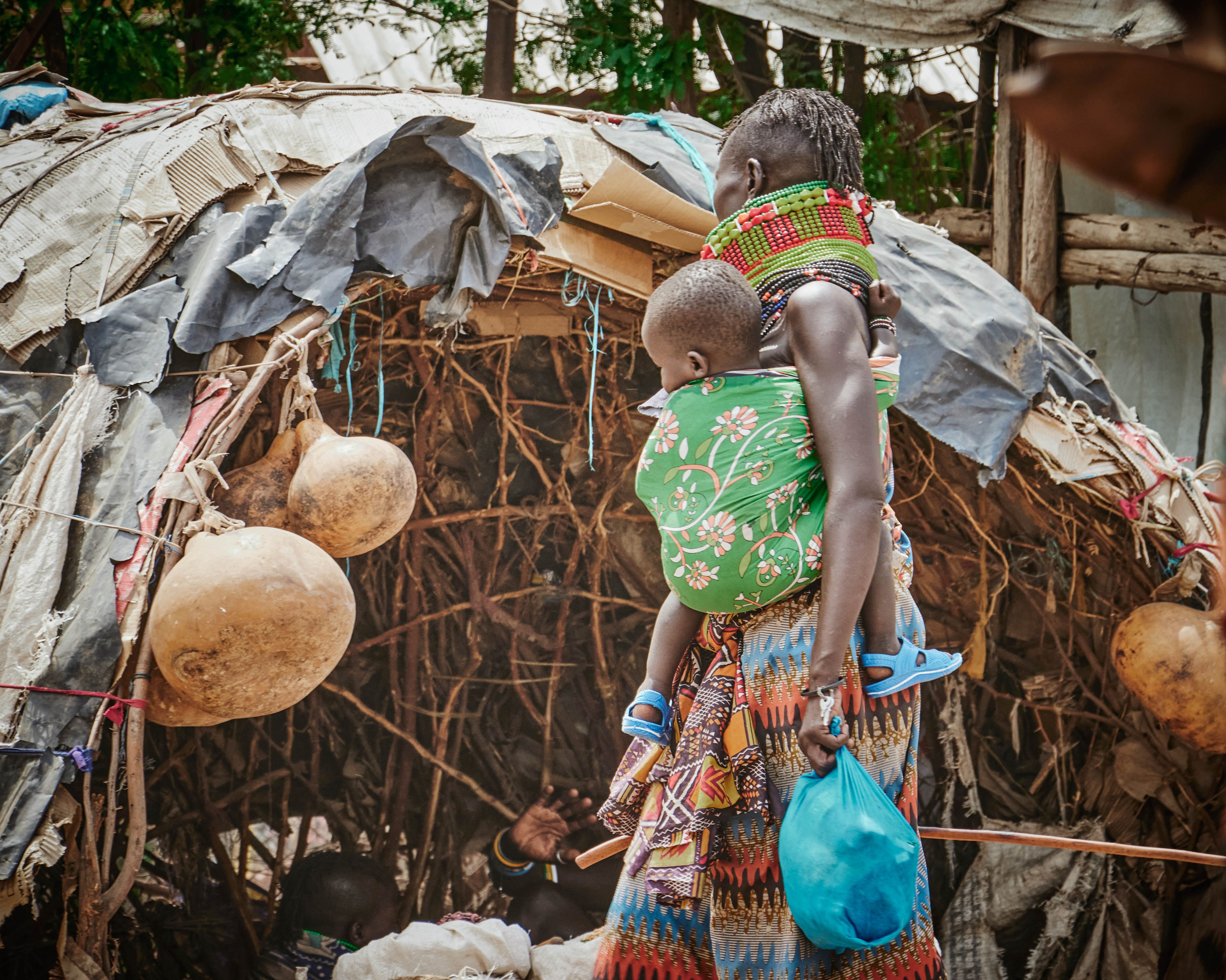 Maasai mother carrying child in vibrant attire near traditional hut in Kenya.