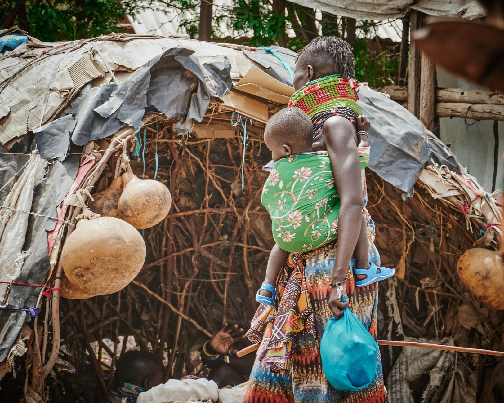 Maasai mother carrying child in vibrant attire near traditional hut in Kenya.