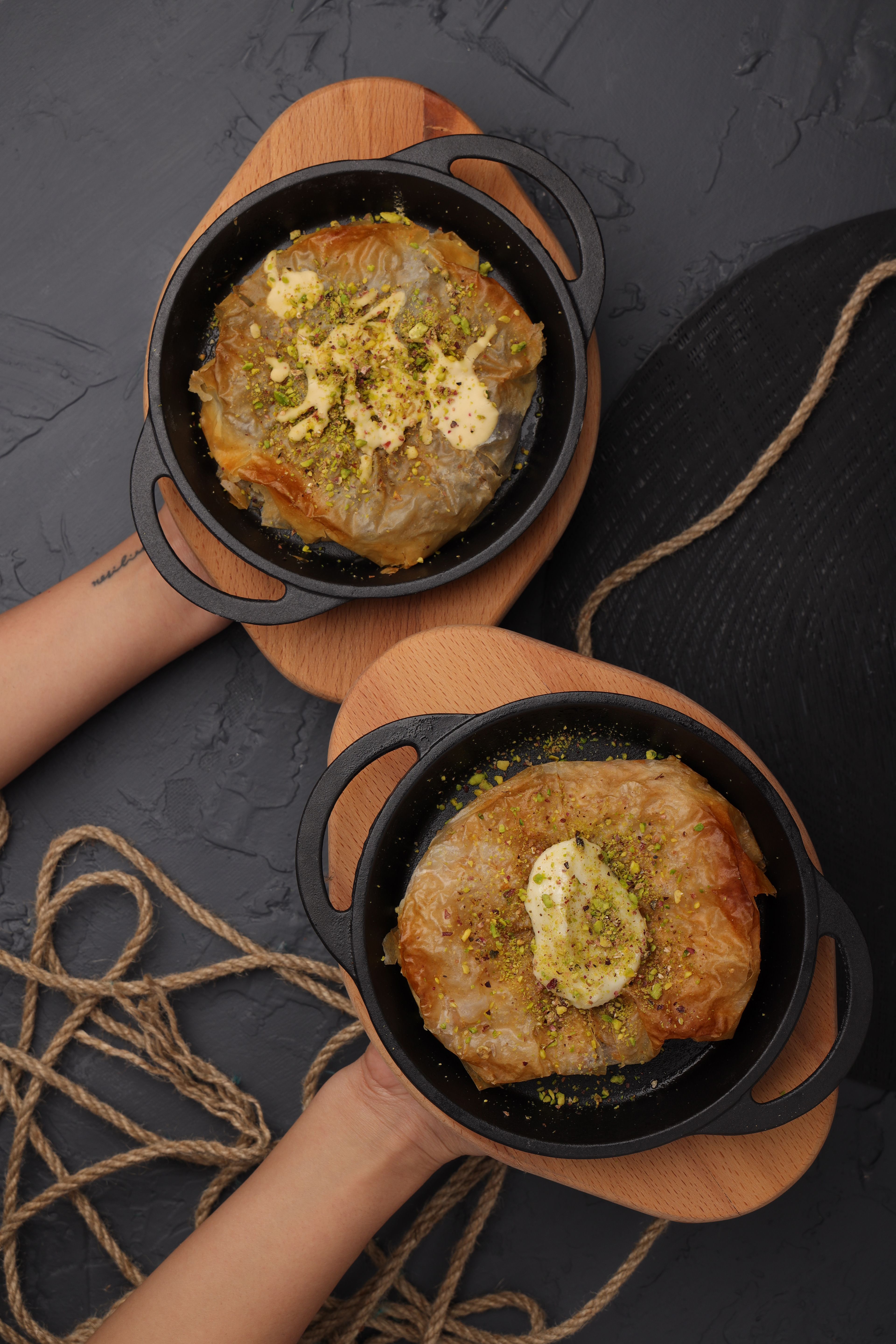 Flat lay of two baklava desserts with pistachios and butter on dark surface.