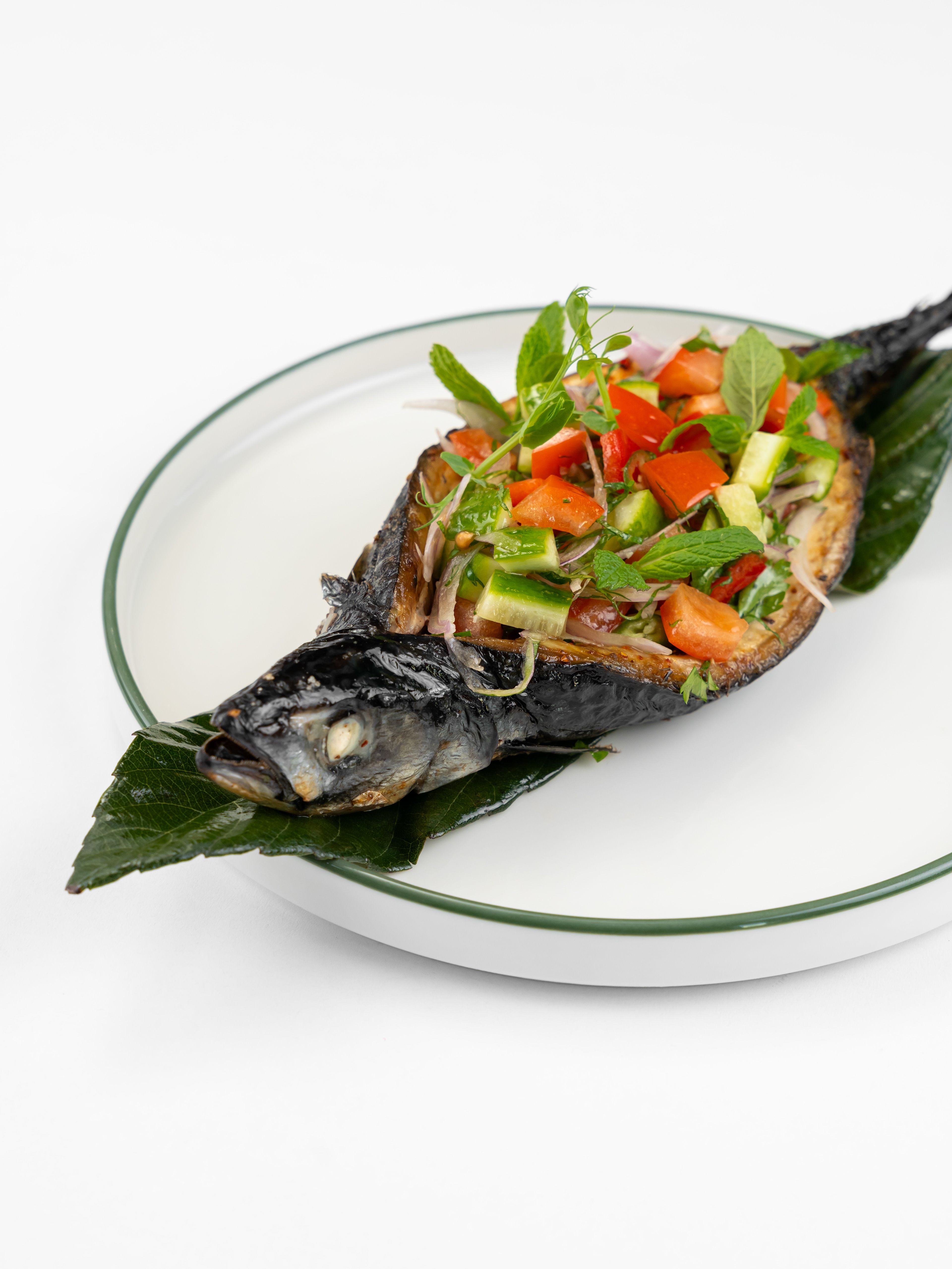 Whole grilled mackerel garnished with fresh tomato and herb salad, served on a leaf-covered plate.