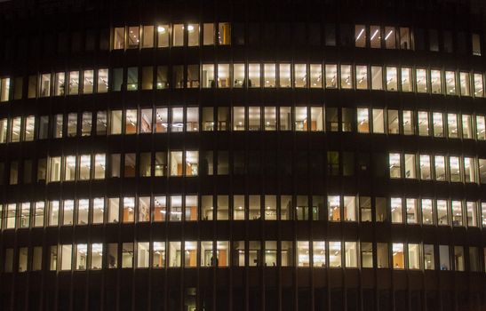 Nighttime view of a brightly lit office building's windows, showcasing interior activity.
