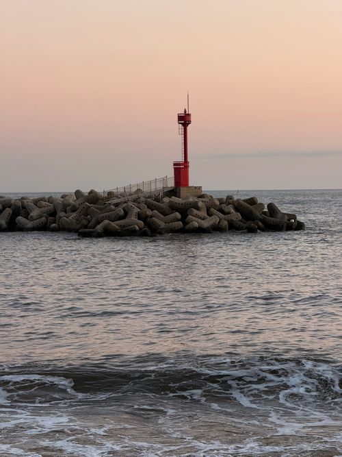 Free A serene view of a red lighthouse on a rocky breakwater, set against a tranquil sea at dusk. Stock Photo