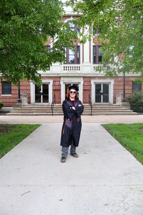 Free Excited graduate wearing academic regalia poses happily in front of a university building. Stock Photo