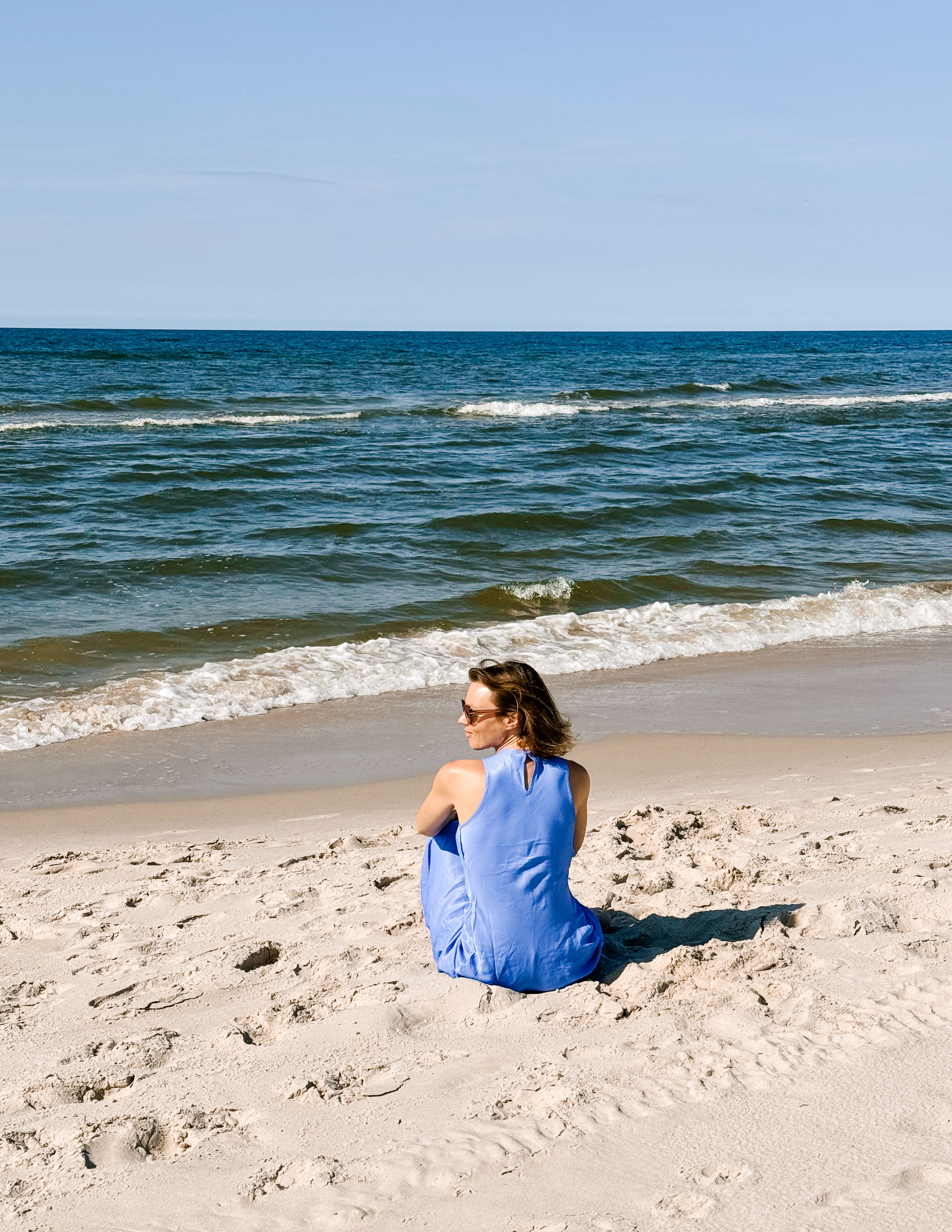 A woman in a blue dress sits on a sandy beach, enjoying a peaceful view of the Baltic Sea.