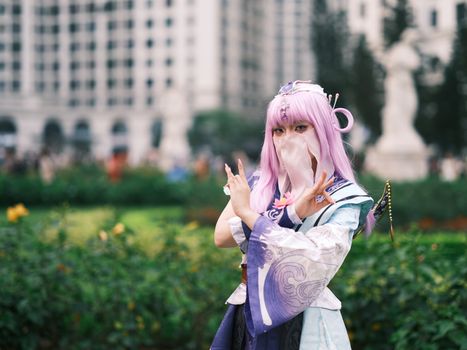 A cosplayer in vibrant costume posing outdoors with a blurred cityscape in the background.